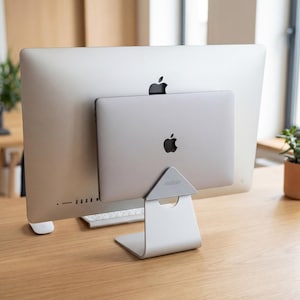 May include: A silver laptop stand holding a silver laptop against the back of a silver computer monitor. The stand has a triangular top and a curved base. The Apple logo is visible on both the laptop and the monitor.