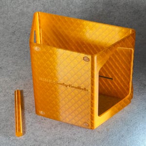 May include: A clear orange plastic box with a diamond pattern on the surface. The box has a small opening on one side and a small hole on the top. The box is sitting on a gray surface. The text "Maker's Key Creations" is printed on the side of the box.