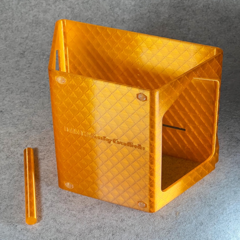 May include: A clear orange plastic box with a diamond pattern on the surface. The box has a small opening on one side and a small hole on the top. The box is sitting on a gray surface. The text "Maker's Key Creations" is printed on the side of the box.