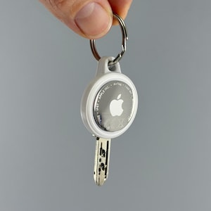 Könnte beinhalten: Ein weißes Apple AirTag an einem Schlüssel mit einem silbernen Schlüsselring befestigt. Das AirTag hat das Apple-Logo und die Aufschrift "AirTag" und "Find My". Der Schlüssel ist silberfarben und hat ein Muster.