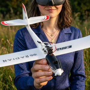 May include: A white and black RC airplane with red wingtip accents is held in front of a person. The plane has the word "RADIAN" printed on the wing. The plane has a camera attached to the front. The person is wearing a blue shirt.