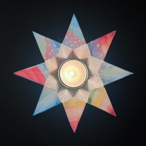 May include: A star-shaped candle holder with a lit candle in the center. The holder is made of folded paper with watercolor patterns in shades of pink, blue, and yellow. The candle holder is on a dark background.