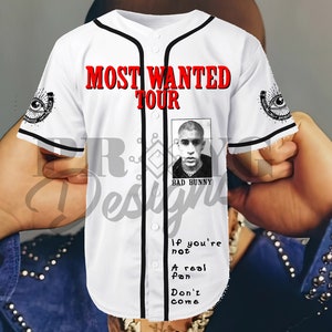 May include: White baseball jersey with black trim and the text "MOST WANTED TOUR" in red. A black and white photo of Bad Bunny is on the front of the jersey with the text "BAD BUNNY" underneath. The text "If you're not a real fan Don't come" is printed on the front of the jersey.
