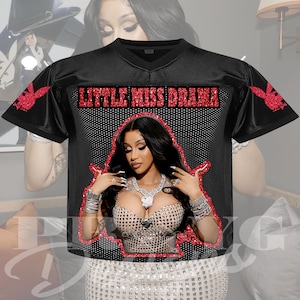 May include: Black football jersey featuring a portrait of a woman with long black hair and jewelry. The jersey has the words "LITTLE MISS DRAMA" in red glitter and a red outline. The sleeves have a red bunny logo.