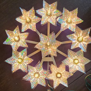 May include: A star-shaped light fixture featuring nine individual six-pointed stars arranged in a circular pattern. Each star has a holographic, iridescent finish and a central light bulb. The stars are mounted on a white frame with a green cord.