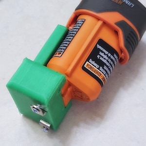May include: A green 3D printed battery holder with two silver screws. The holder is attached to an orange and black battery pack. The battery pack has text in Spanish and English.