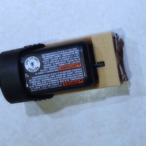 May include: A black and orange battery pack with safety instructions printed on the side. The battery pack is attached to a wooden base with metal clips.