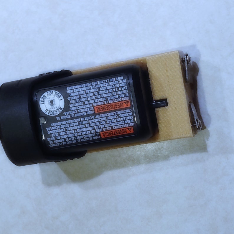 May include: A black and orange battery pack with safety instructions printed on the side. The battery pack is attached to a wooden base with metal clips.