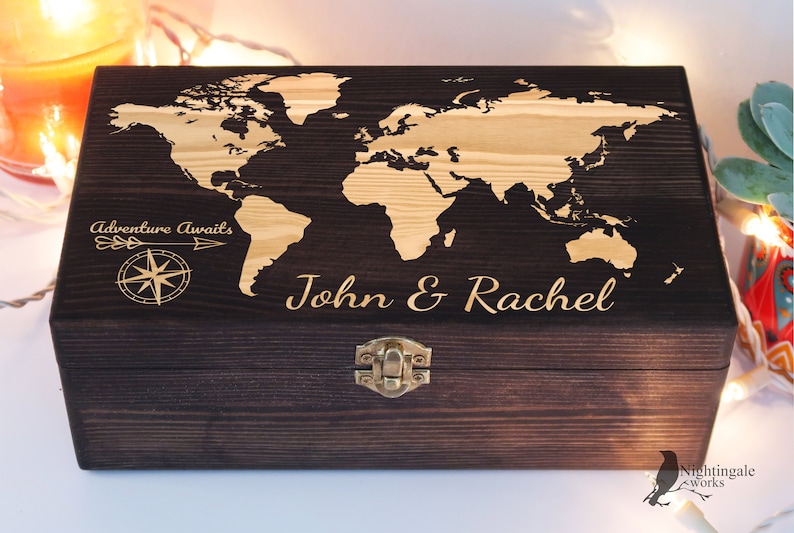 May include: A dark brown wooden box with a world map design engraved on the lid. The map is in a light brown color. The box has a brass latch and is engraved with the text "Adventure Awaits" and "John & Rachel".