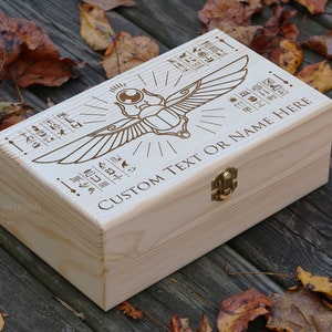 May include: A wooden box with a hinged lid and a brass latch. The box is decorated with an engraved image of a scarab beetle with wings and hieroglyphics. The text "CUSTOM TEXT OR NAME HERE" is engraved on the box.