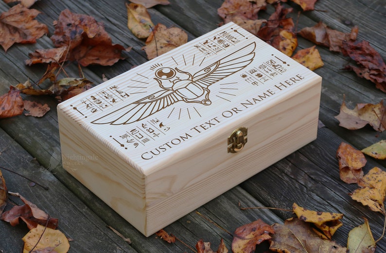 May include: A wooden box with a hinged lid and a brass latch. The box is decorated with an engraved image of a scarab beetle with wings and hieroglyphics. The text "CUSTOM TEXT OR NAME HERE" is engraved on the box.