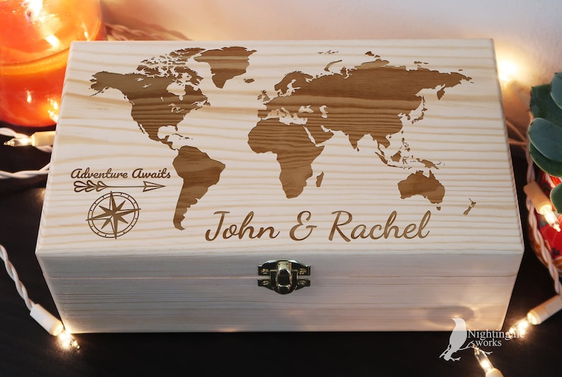 May include: A wooden box with a world map engraved on the lid. The box is light brown and has a brass latch. The text "Adventure Awaits" and a compass are engraved on the left side of the lid. The names "John & Rachel" are engraved on the right side of the lid.