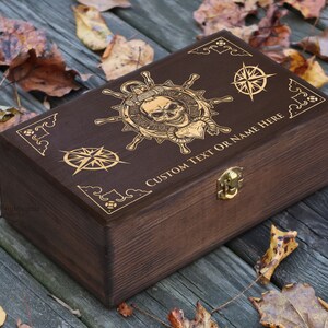 May include: A dark brown wooden box with a gold engraved design of a skull and crossbones, a compass, and the text "CUSTOM TEXT OR NAME HERE".