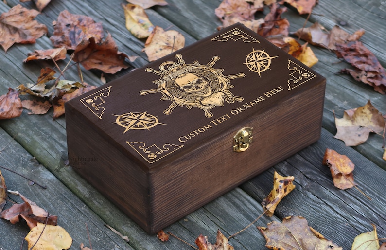 May include: A dark brown wooden box with a gold engraved design of a skull and crossbones, a compass, and the text "CUSTOM TEXT OR NAME HERE".