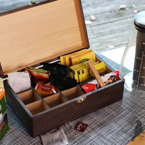 May include: A wooden tea box with a hinged lid, containing various tea bags in individual compartments. The box is open and the lid is propped up. There is a glass tea pitcher with a stainless steel lid and a measuring scale on the side. The tea box and pitcher are on a gray placemat.