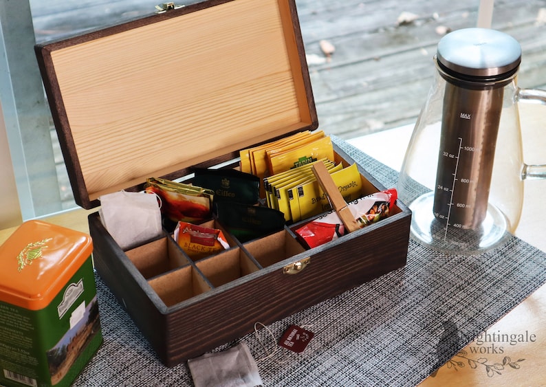 May include: A wooden tea box with a hinged lid, containing various tea bags in individual compartments. The box is open and the lid is propped up. There is a glass tea pitcher with a stainless steel lid and a measuring scale on the side. The tea box and pitcher are on a gray placemat.