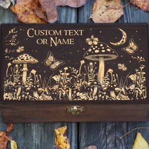 Pode incluir: Uma caixa retangular de madeira castanho-escura com um fecho de metal dourado. A tampa apresenta um design gravado de cogumelos, borboletas e uma lua crescente, com o texto CUSTOM TEXT OR NAME centrado no topo da superfície da tampa.