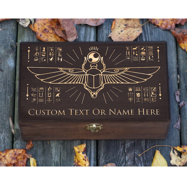 May include: A dark brown wooden box with a gold engraved design of a scarab beetle with wings and hieroglyphics. The box has a hinged lid and a brass latch. The text "CUSTOM TEXT OR NAME HERE" is engraved on the front of the box.