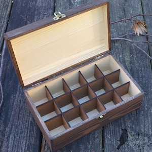 May include: A wooden box with a hinged lid and 16 compartments. The box is made of dark wood with a light wood interior. The box is open and the compartments are empty.