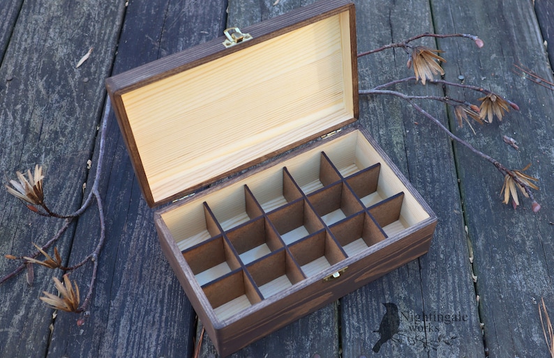 May include: A wooden box with a hinged lid and 16 compartments. The box is made of dark wood with a light wood interior. The box is open and the compartments are empty.