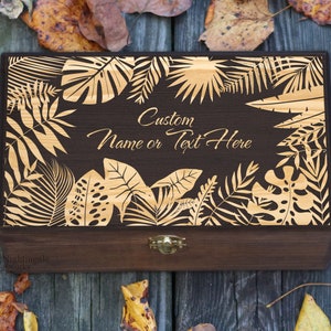 May include: A dark brown wooden box with a gold-colored tropical leaf design and a hinged lid. The box has a brass latch and the text "Custom Name or Text Here" is engraved on the front.