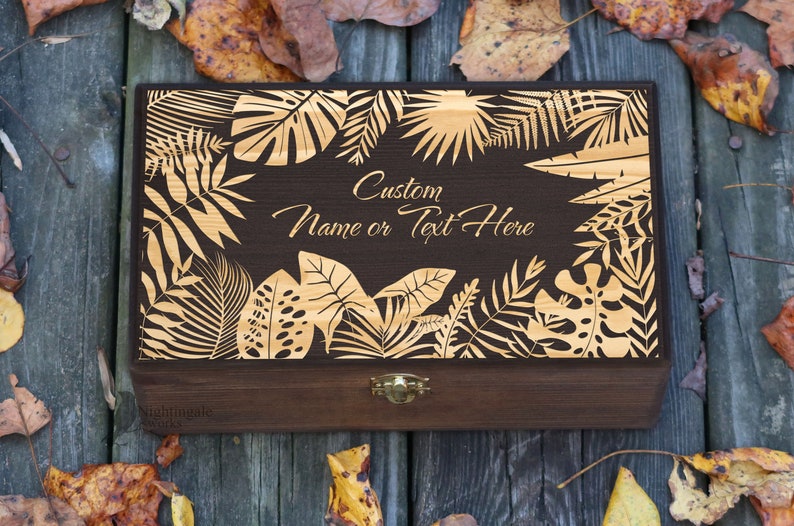 May include: A dark brown wooden box with a gold-colored tropical leaf design and a hinged lid. The box has a brass latch and the text "Custom Name or Text Here" is engraved on the front.