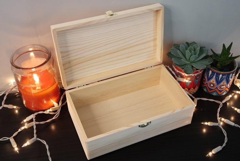 May include: A wooden box with a hinged lid, open to reveal the empty interior. The box is made of natural wood with a light brown finish. The box is sitting on a dark surface with string lights and potted plants.