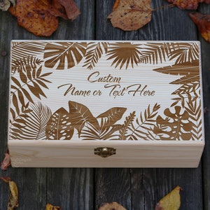 May include: A wooden box with a hinged lid and a brass latch. The lid is decorated with a laser-cut design of tropical leaves. The text "Custom Name or Text Here" is engraved on the lid.
