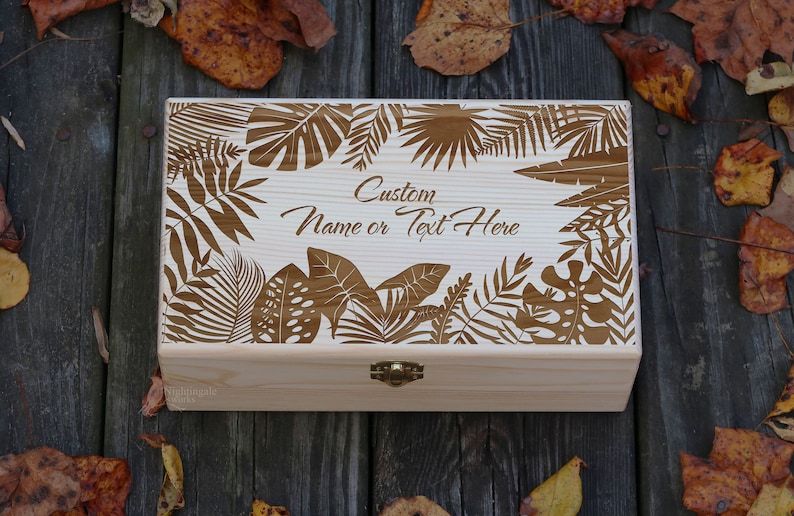 May include: A wooden box with a hinged lid and a brass latch. The lid is decorated with a laser-cut design of tropical leaves. The text "Custom Name or Text Here" is engraved on the lid.