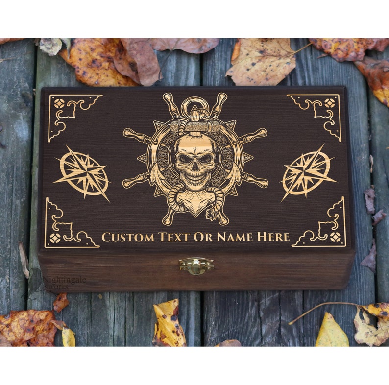 May include: A wooden box with a dark brown finish and a gold engraved design of a skull with a nautical theme. The box has a hinged lid and a brass latch. The text "CUSTOM TEXT OR NAME HERE" is engraved on the front of the box.