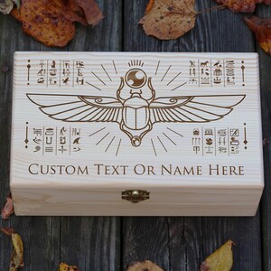 May include: A wooden box with a hinged lid, featuring an engraved design of a scarab beetle with wings and hieroglyphics. The box has a brass latch and the text "CUSTOM TEXT OR NAME HERE" is engraved on the front.