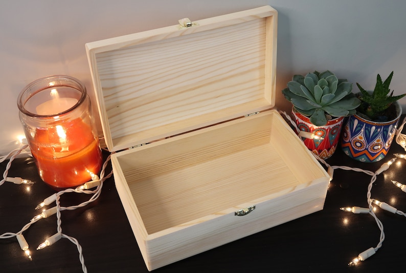 May include: A natural wood box with a hinged lid and brass clasp. The box is open and empty, revealing the natural wood grain inside. The box is sitting on a dark surface with string lights and potted succulents.