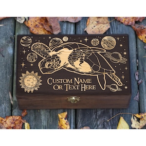 May include: A dark brown wooden box with an engraved design of a sea turtle, planets, stars, and a sun. The text "CUSTOM NAME OR TEXT HERE" is engraved on the box. It has a brass-coloured clasp. The box is suitable for storing small items.