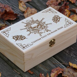 May include: A wooden box with a hinged lid and a brass latch. The box is decorated with an engraved design of a skull and crossbones, a compass, and the text "CUSTOM TEXT OR NAME HERE".