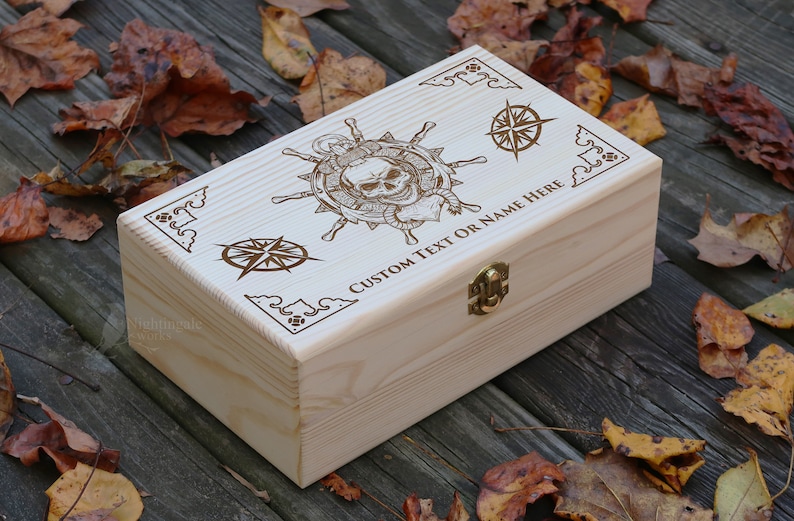 May include: A wooden box with a hinged lid and a brass latch. The box is decorated with an engraved design of a skull and crossbones, a compass, and the text "CUSTOM TEXT OR NAME HERE".