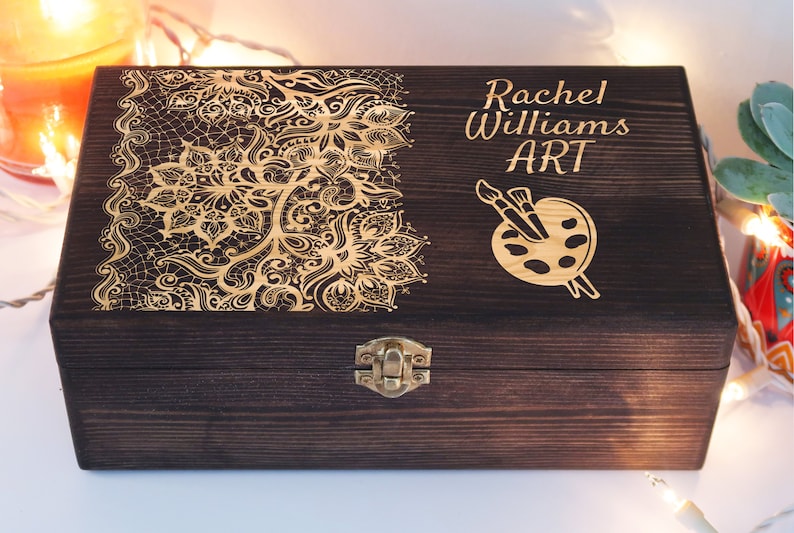 May include: A dark brown wooden box with a gold engraved design on the lid. The design features a floral pattern and a paint palette with paintbrushes. The box is engraved with the text "Rachel Williams ART".
