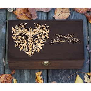 May include: A dark brown wooden box with a gold-colored floral design and a medical symbol. The box is engraved with the name "Meredith Johnson M.D." and has a small gold clasp. The box is surrounded by fall leaves.