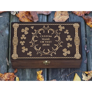 Puede incluir: Una caja de madera marrón oscuro con diseños dorados de nudos celtas y tréboles. La caja presenta el texto "CUSTOM NAME OR TEXT HERE" en un marco decorativo. Un pequeño cierre dorado asegura la caja.