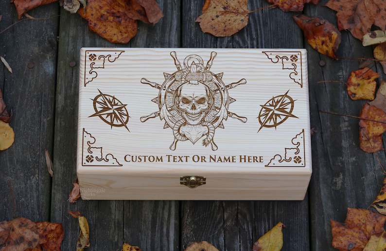 May include: A wooden box featuring a skull and crossbones design, a ship's wheel, and compasses. The box has a hinged lid and a brass latch. The text "CUSTOM TEXT OR NAME HERE" is engraved on the front of the box.
