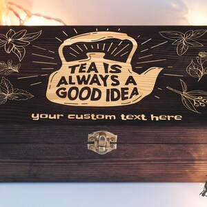 May include: A dark brown wooden tea box with a gold engraved design of a teapot and the text "Tea is always a good idea". The box has a hinged lid with a brass clasp.