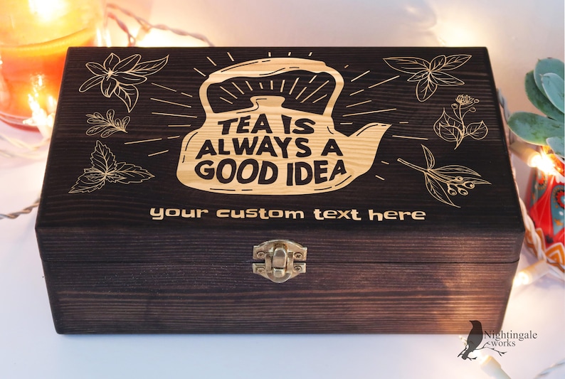 May include: A dark brown wooden tea box with a gold engraved design of a teapot and the text "Tea is always a good idea". The box has a hinged lid with a brass clasp.