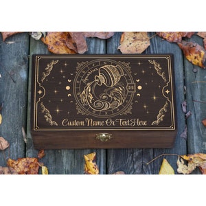 May include: A dark brown wooden box with a gold engraved design of the zodiac sign Aquarius. The box has a brass latch and the text "Custom Name Or Text Here" is engraved on the front.
