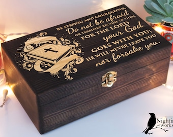 Puede incluir: Una caja de madera oscura con un cierre dorado y un diseño de biblia en la tapa. La caja tiene una inscripción grabada que dice: "Sé fuerte y valiente. No tengas miedo ni te aterrorices por causa de ellos. Porque el Señor tu Dios va contigo; él nunca te dejará ni te abandonará."