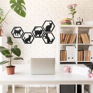 Pode incluir: Silhuetas de gatos pretos de metal em molduras hexagonais montadas em uma parede de tijolo branco. A decora&ccedil;&atilde;o de parede &eacute; exibida em um ambiente de escrit&oacute;rio em casa com uma mesa branca, um laptop, uma planta em vaso e uma estante.