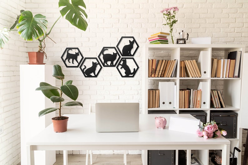 Pode incluir: Silhuetas de gatos pretos de metal em molduras hexagonais montadas em uma parede de tijolo branco. A decora&ccedil;&atilde;o de parede &eacute; exibida em um ambiente de escrit&oacute;rio em casa com uma mesa branca, um laptop, uma planta em vaso e uma estante.