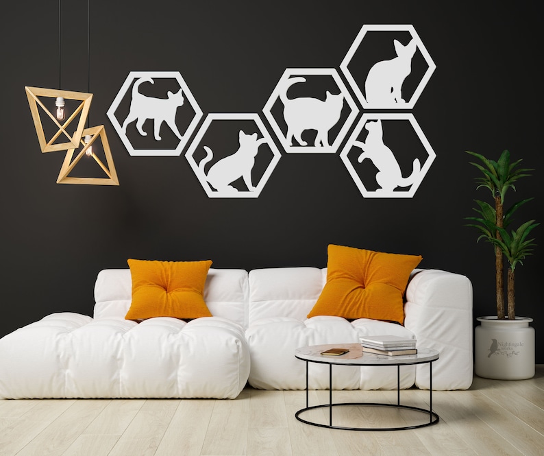 Pode incluir: Silhuetas brancas de gatos em formas hexagonais s&atilde;o dispostas em uma parede preta. A decora&ccedil;&atilde;o da parede est&aacute; acima de um sof&aacute; branco com duas almofadas laranja. Uma mesa de centro redonda com uma moldura de metal preto fica na frente do sof&aacute;.