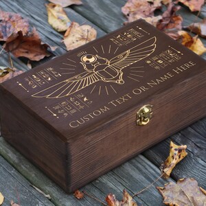 May include: A dark brown wooden box with a gold engraved design of an Egyptian scarab beetle with wings. The box has a hinged lid and a brass latch. The text "CUSTOM TEXT OR NAME HERE" is engraved on the box.