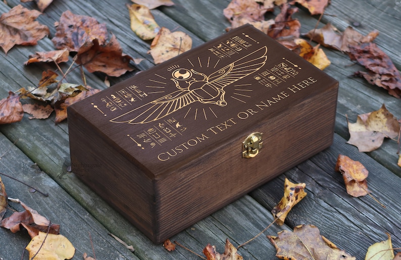 May include: A dark brown wooden box with a gold engraved design of an Egyptian scarab beetle with wings. The box has a hinged lid and a brass latch. The text "CUSTOM TEXT OR NAME HERE" is engraved on the box.