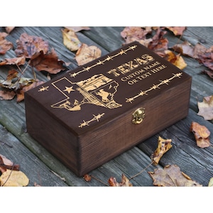 May include: A dark brown wooden box featuring a gold-coloured Texas design and the text "TEXAS CUSTOM NAME OR TEXT HERE". The box has a gold-coloured clasp and barbed wire detailing.