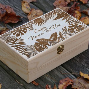 May include: A wooden box with a hinged lid and a brass latch. The lid is decorated with a laser-cut design of tropical leaves and the text "Custom Name or Text Here".